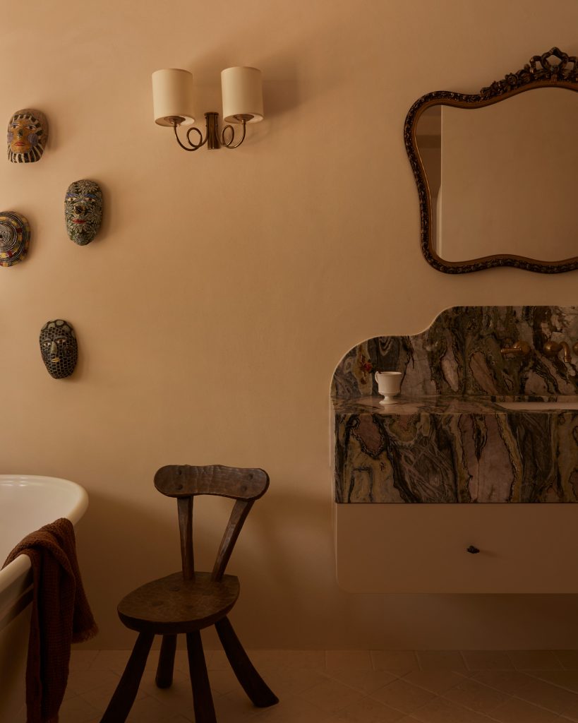 Bathroom with beige walls, featuring a white bathtub with a brown towel, four decorative masks on the wall, a rustic wooden chair, a sink with a marble countertop, an ornate mirror, and a wall-mounted light fixture