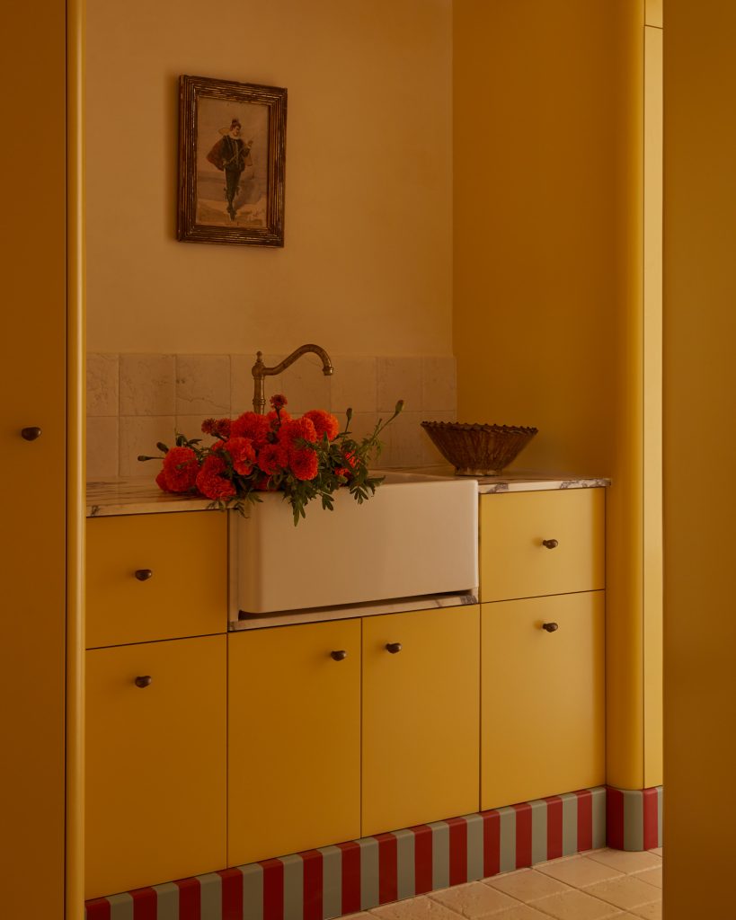 Kitchen area with yellow cabinets and drawers, a white sink with a brass faucet, light-colored tile backsplash, a bouquet of red flowers in a vase, a decorative bowl on the countertop, and a framed picture above the backsplash.