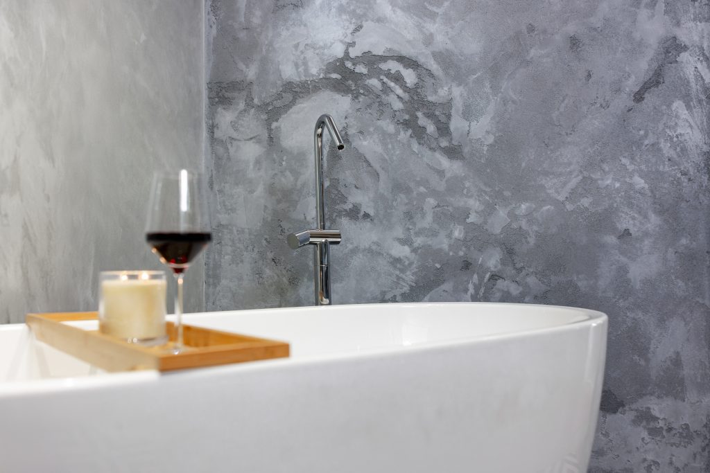 Modern bathroom with Rockcote MicroStone walls, featuring a freestanding white bathtub, a wooden ladder holding towels, hanging plants, and geometric black-and-white floor tiles.