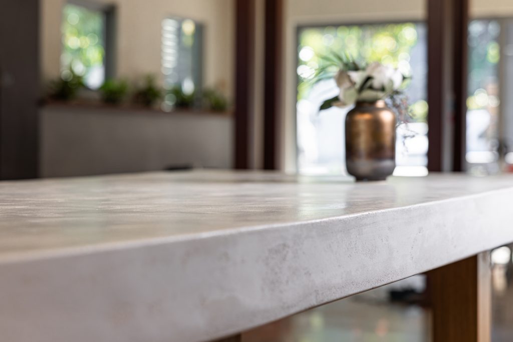 Close-up view of a smooth, light-colored countertop with a blurred vase of white flowers and greenery in the background, set indoors with large windows and visible plants.