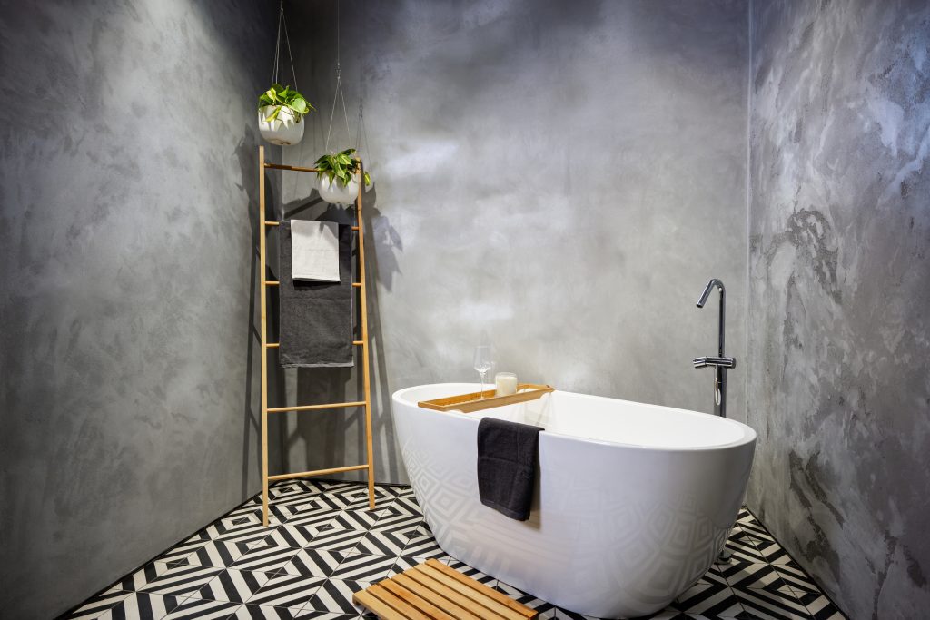 Modern bathroom with Rockcote MicroStone walls, featuring a freestanding white bathtub, a wooden ladder holding towels, hanging plants, and geometric black-and-white floor tiles.